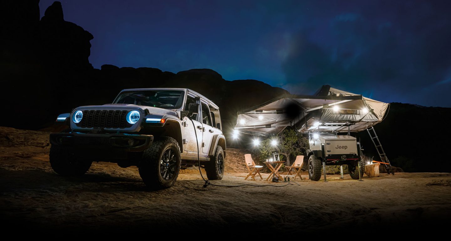 Un Jeep 4xe 2024 estacionado junto a un campamento por la noche, con un extremo de un cable inversor conectado al puerto de carga del vehículo y el otro enchufado al equipo de campamento. El camping dispone de una autocaravana marca Jeep equipada con toldos y luces.