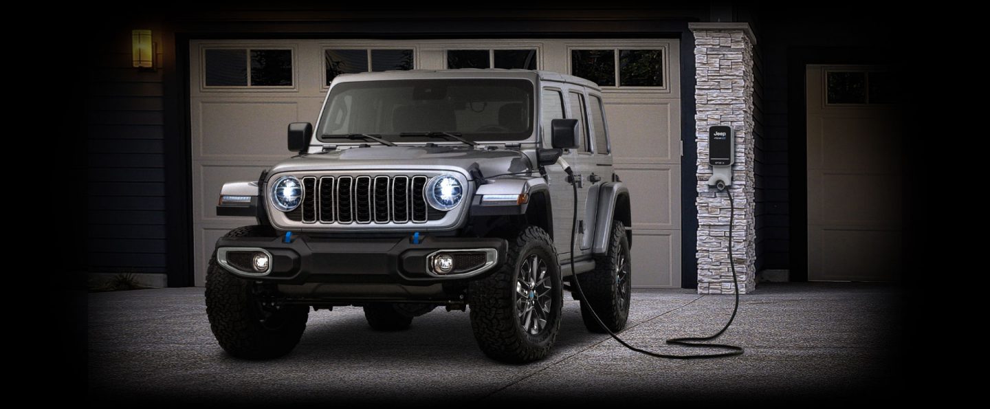 Un Jeep Wrangler Sahara 4xe 2024 estacionado frente a un garaje con una estación de carga incorporada y un cable enchufado en el puerto de carga del vehículo.